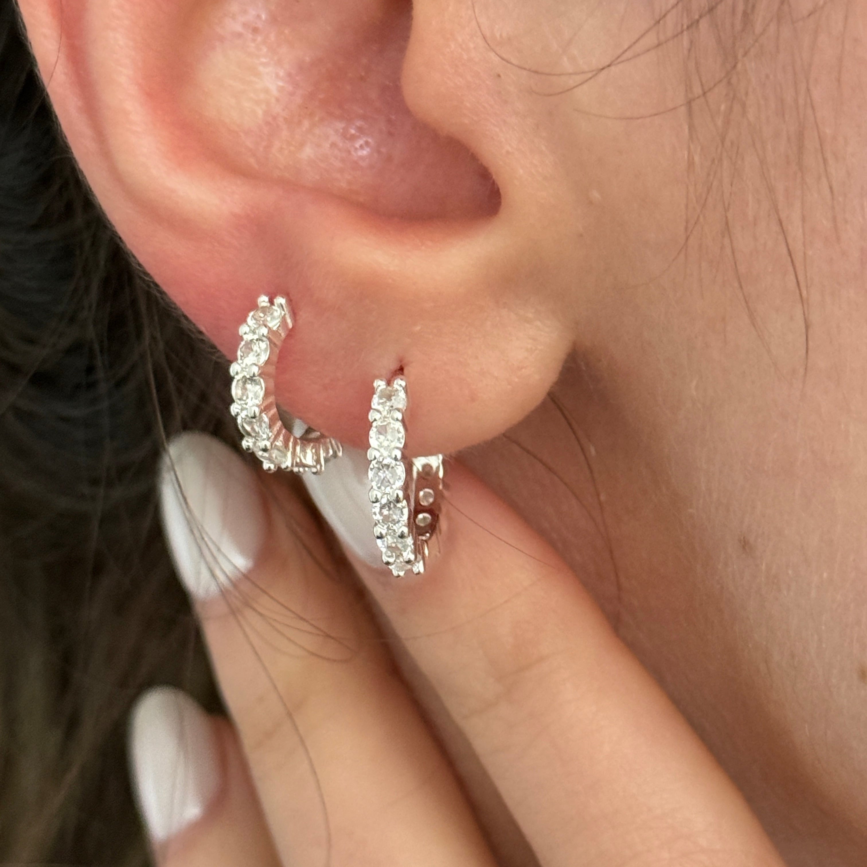 A close-up of a person's ear wearing two small silver huggie hoop earrings set with clear stones, with fingers touching the ear.