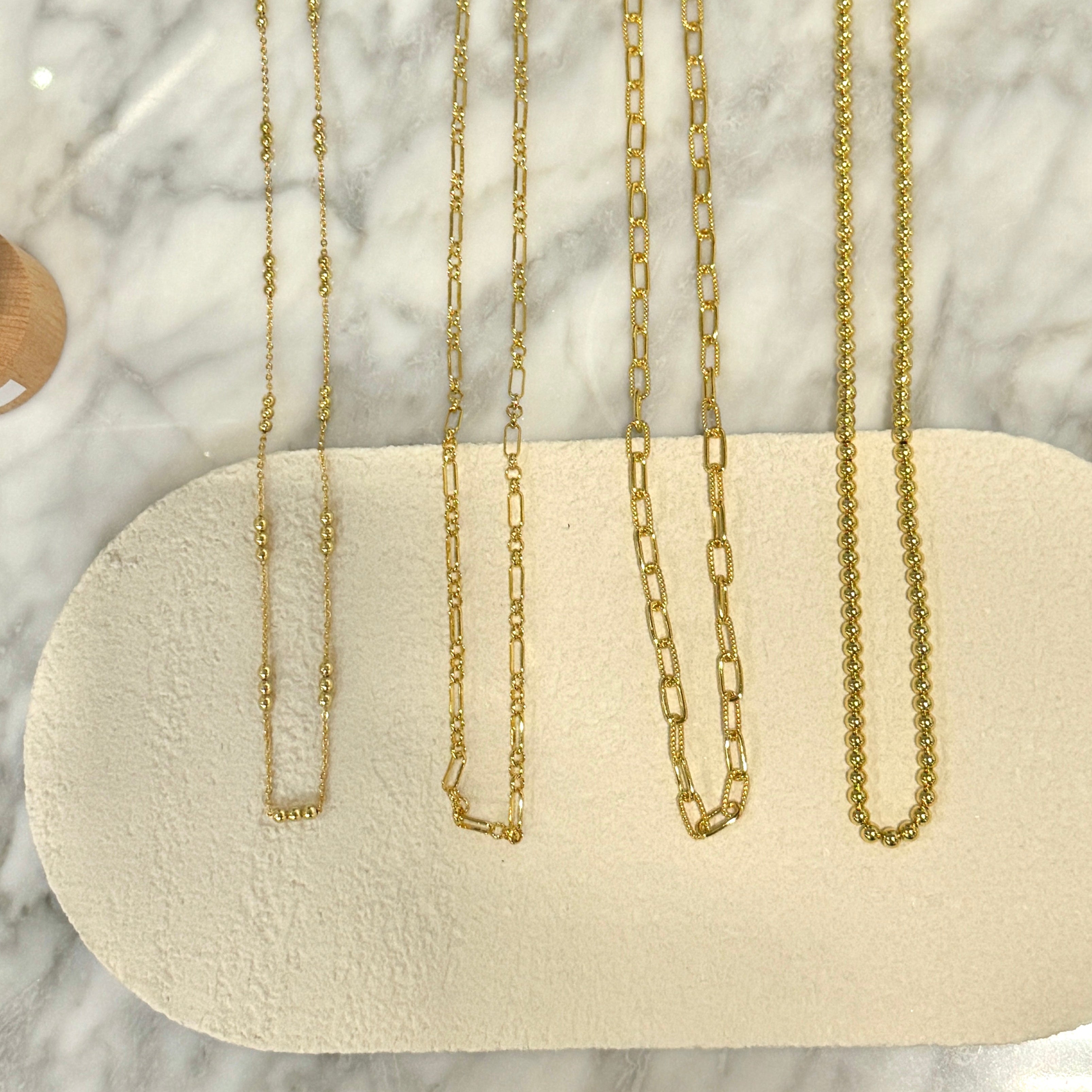 Four gold-toned chain necklaces in different link styles are displayed on a beige oval jewelry stand over a white marble surface. Small white tags are attached to each necklace, and part of a tablet is visible at the top of the frame.