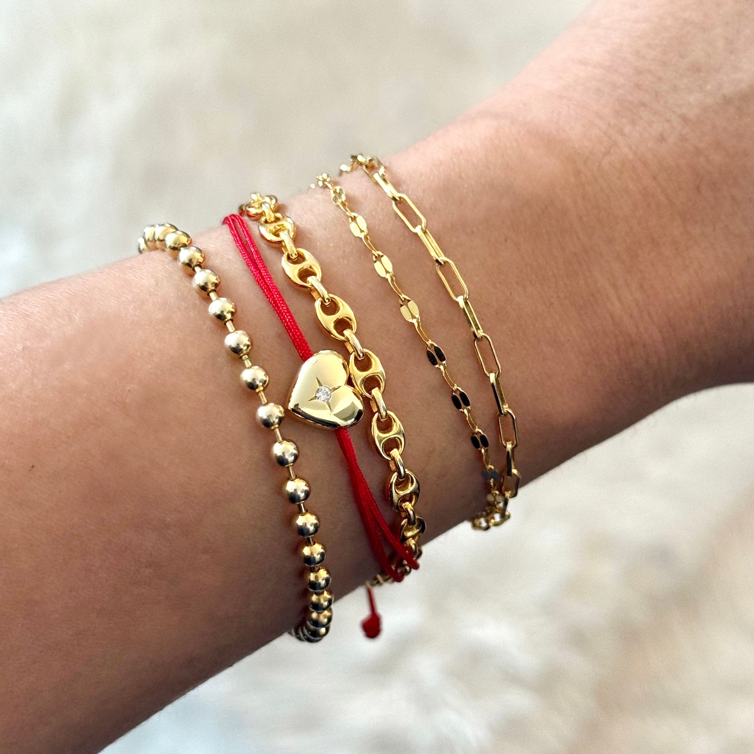 Gold heart charm red string bracelet stacked with other delicate bracelets on model’s wrist