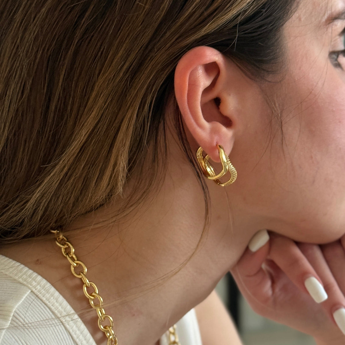 A woman in profile wearing a gold chain necklace and matching gold hoop earrings, resting her chin on her hand in an indoor setting.