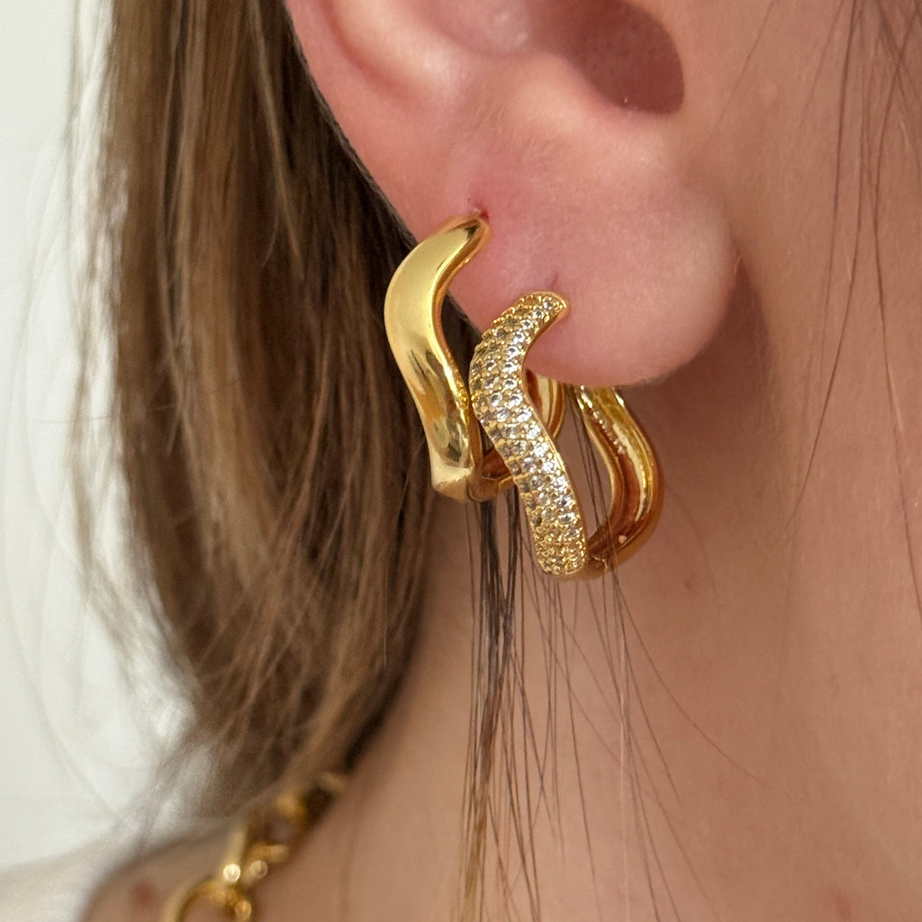 A close-up of a person's ear wearing a pair of gold huggie hoop earrings, one plain gold and the other covered in small clear pavé stones.
