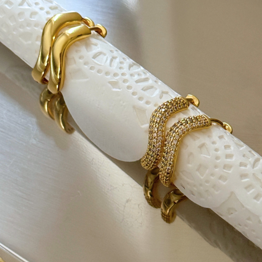 Two gold wrap-style hoop earrings displayed on a carved white cylindrical stand. One earring is smooth polished gold, and the other is covered in small clear stones.